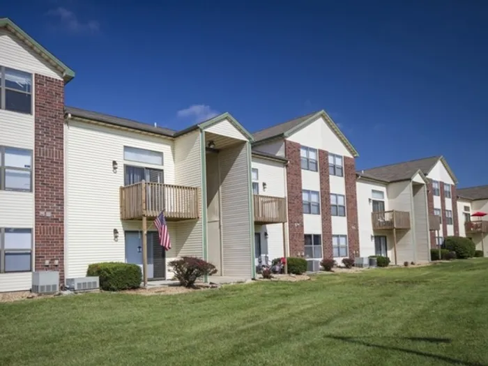 Three-story apartment buildings with beige siding and red brick accents, private balconies, and large windows, set against a bright blue sky with a wide, well-maintained green lawn in front.
