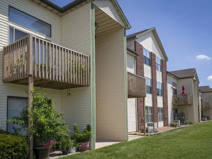 Side angle of the apartment building featuring beige siding, brick accents, and private balconies&mdash;some with potted plants&mdash;overlooking a green lawn.
