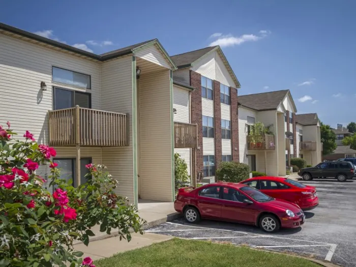 Exterior view showing the apartments&rsquo; architecture and on-site parking area with red cars, framed by blooming rose bushes and a bright blue sky.