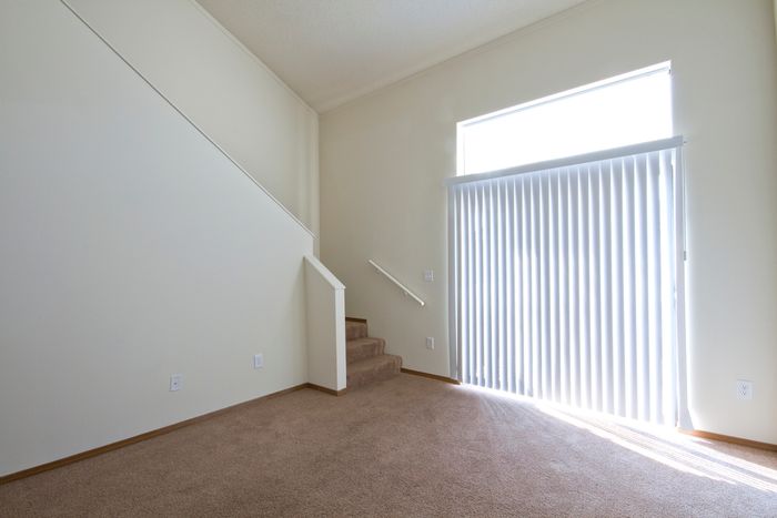 Bright living space with high ceilings, vertical blinds covering a sliding glass door, and carpeted stairs leading to an upper level.