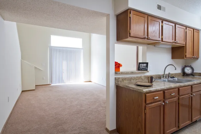 Open floor plan connecting the kitchen to a carpeted living area with natural light pouring in through vertical blinds and access to a private balcony.