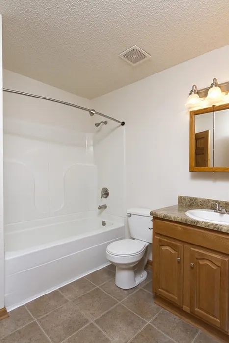 Simple and clean bathroom layout with full tub and shower combo, wood vanity, granite-look countertop, and a mounted mirror with light fixture.