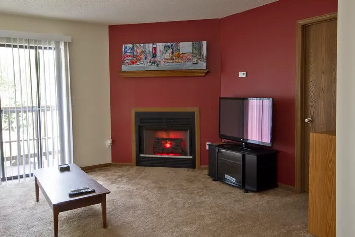Cozy living space with a corner fireplace set against a deep red accent wall, carpet flooring, mounted artwork, and a TV setup near a sliding glass door.