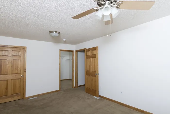 Standard bedroom layout with neutral carpet, ceiling fan, light fixture, and access to a hallway and closet with wooden doors.