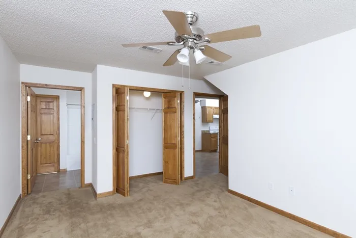 Spacious bedroom featuring two open closets with bi-fold wooden doors, ceiling fan, and view into an adjacent kitchen and hallway.