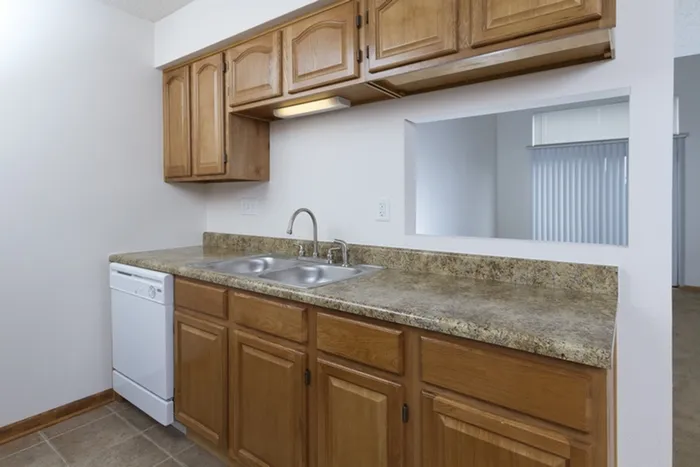 Functional kitchen with natural wood cabinets, granite-look countertops, a double sink, dishwasher, and a passthrough window into the living area.