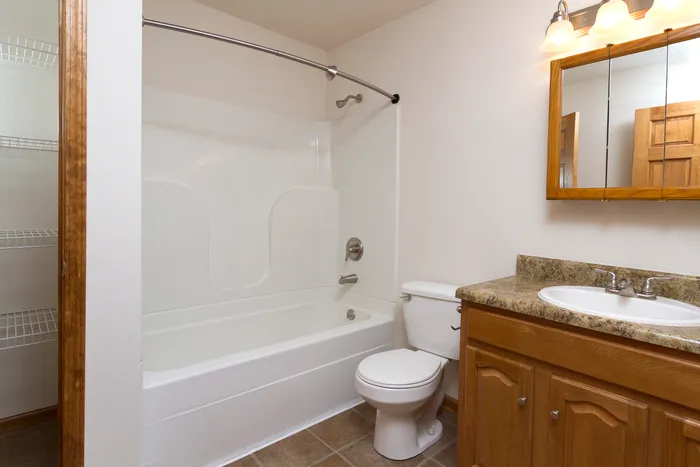 Clean and simple bathroom with a white shower/tub combo, wood vanity, granite-style countertop, toilet, and a framed mirror with lighting.