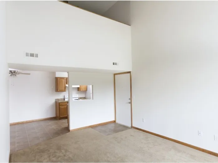 Empty room with high ceiling and white walls. A small kitchen area with wooden cabinets is visible. The space is minimalistic and bright.