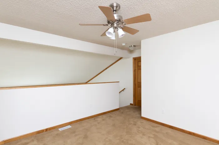 A small, empty room with beige carpet and white walls; a wooden railing lines a staircase. A ceiling fan with wooden blades is centered above.