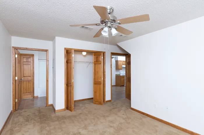 Empty room with beige carpet, white walls, and a ceiling fan with wooden blades. Two open wooden doors lead to a closet and another room, creating a spacious feel.