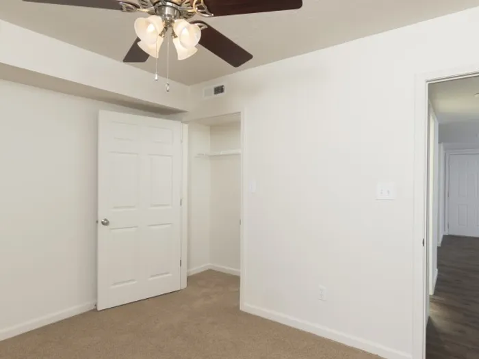 Bedroom with soft carpeting, neutral walls, a ceiling fan with light fixture, an open closet with shelving, and a door leading to a hallway in a modern apartment.