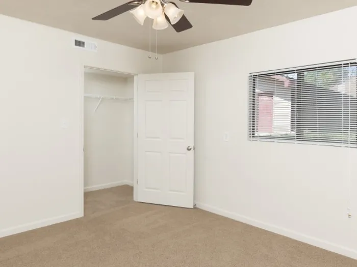 Bright bedroom with soft carpeting, neutral walls, a ceiling fan with light fixture, an open closet, and a window with blinds offering natural light in a modern apartment.