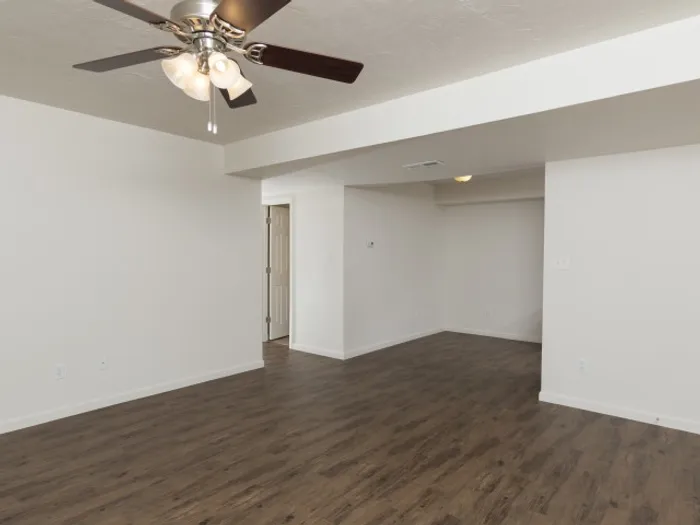Spacious living area with hardwood-style flooring, a ceiling fan with light fixtures, and neutral-colored walls, providing a versatile space in a modern apartment unit