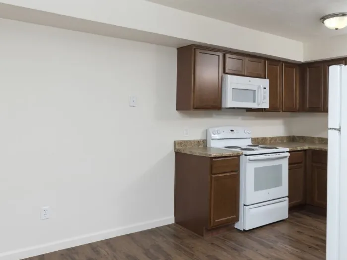 Kitchen area with wood cabinetry, white appliances including a stove and microwave, granite-style countertops, and hardwood-style flooring in a modern apartment unit.