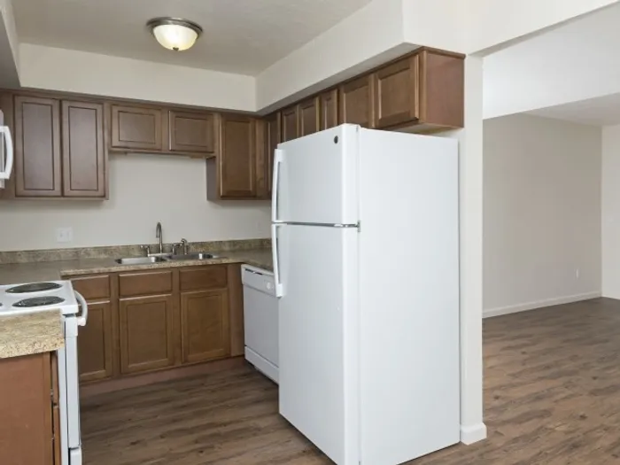 Modern kitchen with wood cabinetry, white appliances including a refrigerator and dishwasher, granite-style countertops, and hardwood-style flooring, leading to an open living space in a professionally managed apartment community.