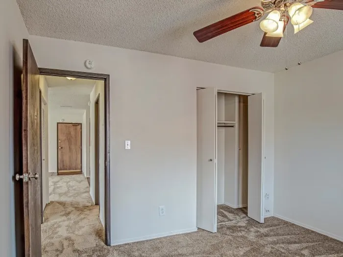 Bedroom with plush carpeting, neutral walls, a ceiling fan with light fixture, an open closet, and a view of the hallway leading to other rooms in a modern apartment.