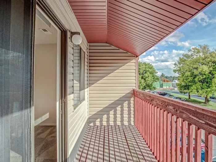 Private balcony with red railing and siding, featuring an open view of greenery and a parking area, accessible from a sliding glass door in a modern apartment.