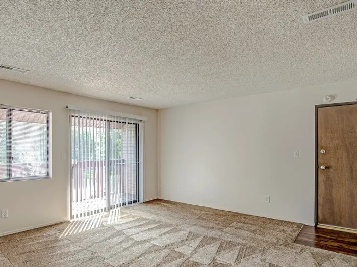 Bright living room with textured carpeting, large windows, and sliding glass doors leading to a private balcony, creating a welcoming and airy space in a modern apartment unit.