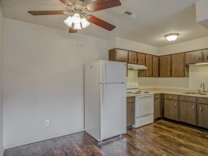 Cozy kitchen with wood-style cabinetry, white appliances including a refrigerator and stove, hardwood-style flooring, and a ceiling fan with light fixture, providing a functional and inviting space in a modern apartment.