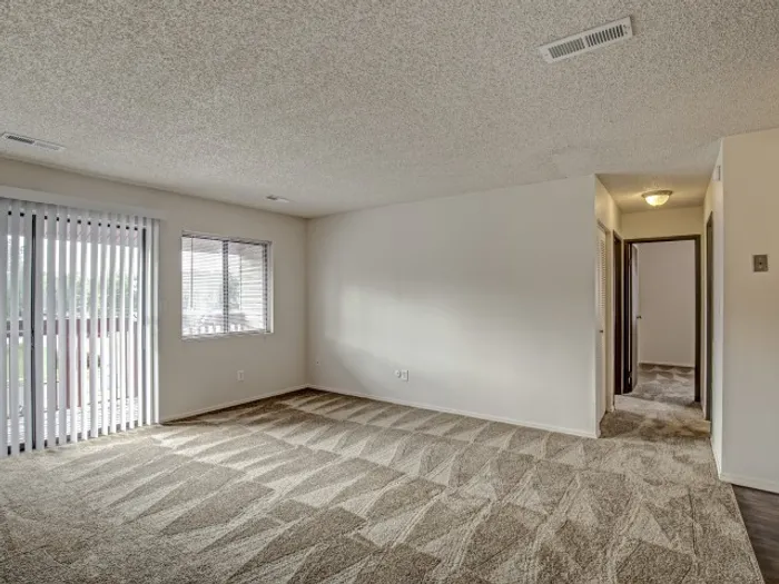 Spacious living room with freshly cleaned textured carpeting, large windows, sliding glass doors to a private balcony, and a hallway leading to additional rooms, offering a comfortable and functional layout in a modern apartment.