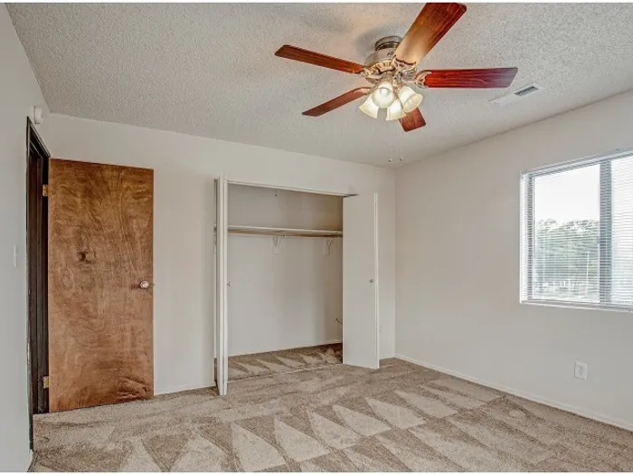 Bright bedroom with textured carpeting, a ceiling fan with light fixture, a large window for natural light, and an open closet with ample storage space in a modern apartment.