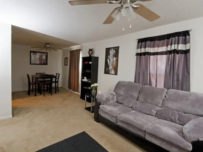 Cozy living room with gray sofa, beige carpet, and a ceiling fan. Dining table in the background. Warm, inviting atmosphere with soft lighting.