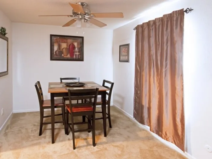 A cozy dining room with a dark wooden table and four chairs. A ceiling fan hangs above. Light filters through brown curtains, creating a warm, inviting atmosphere.