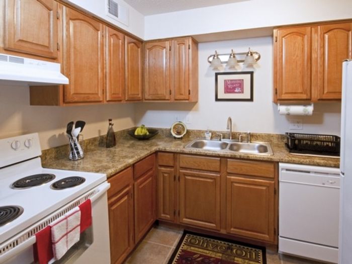 A cozy kitchen with wooden cabinets, a light countertop, and white appliances. A decorative wall piece and a fruit bowl add warmth and charm.