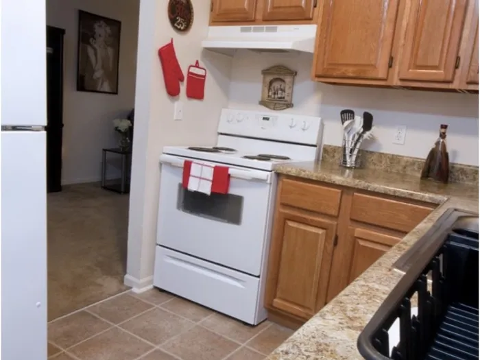 Cozy kitchen with wooden cabinets, a white stove adorned with red checkered towels. Warm lighting and tile floors create a welcoming atmosphere.
