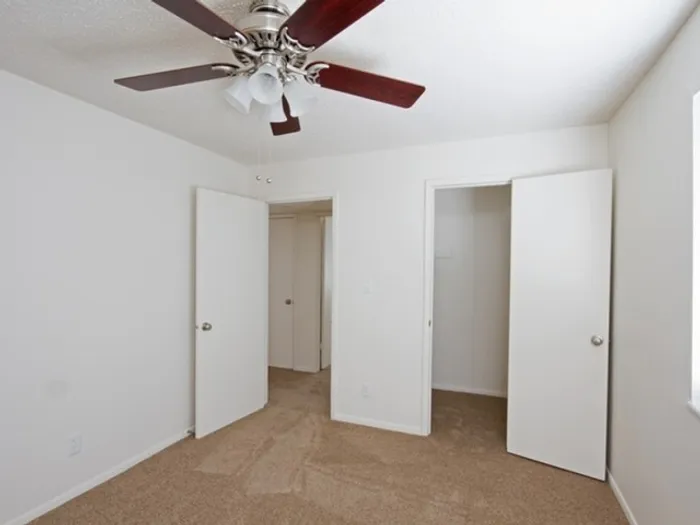 A white, empty room with beige carpet, featuring a ceiling fan with wooden blades. Open doors lead to a hallway and a closet, creating a simple, clean look.
