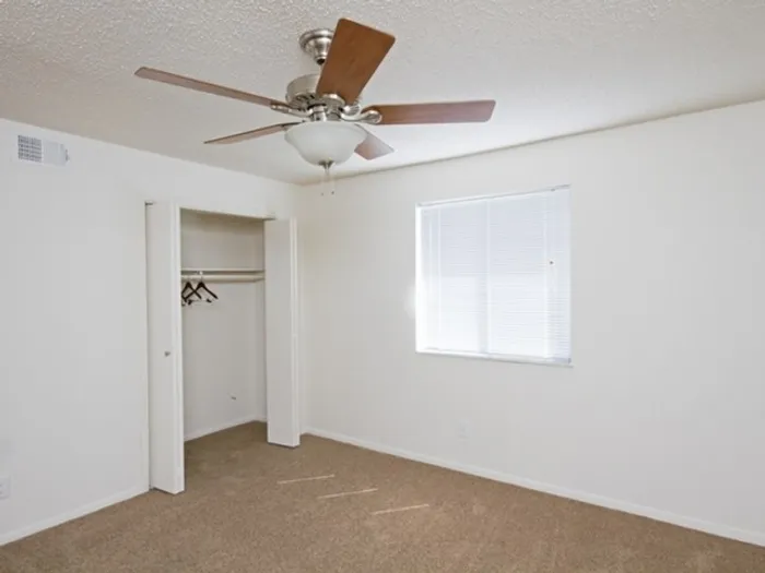 Empty room with white walls and beige carpet, featuring an open closet with hangers, a large window with blinds, and a ceiling fan creating a minimalist feel.