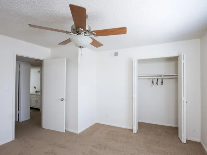 Empty white room with carpet flooring, open closet, ceiling fan, and two doors leading to a kitchen area and another space. Minimalist and clean.