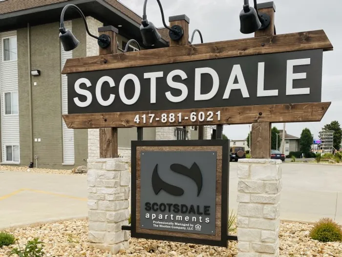 Sign for Scotsdale Apartments with a phone number, mounted on stone pillars. The backdrop includes a building and overcast sky, conveying a calm, residential atmosphere.