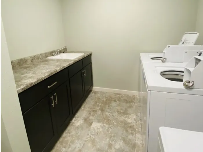 Laundry room with a white washer and dryer on the right, dark wood cabinets with a marble countertop on the left, and light beige walls and flooring.