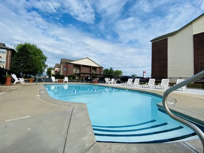 Large outdoor pool with a curved edge, surrounded by white lounge chairs and apartment buildings, under a partly cloudy sky.