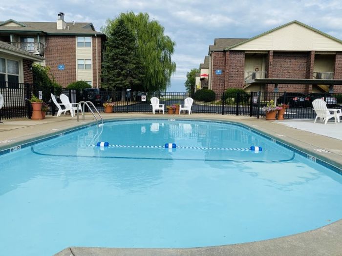 Clear pool water with a roped-off swim section, white patio chairs, and apartments lining the background on a partly cloudy day.