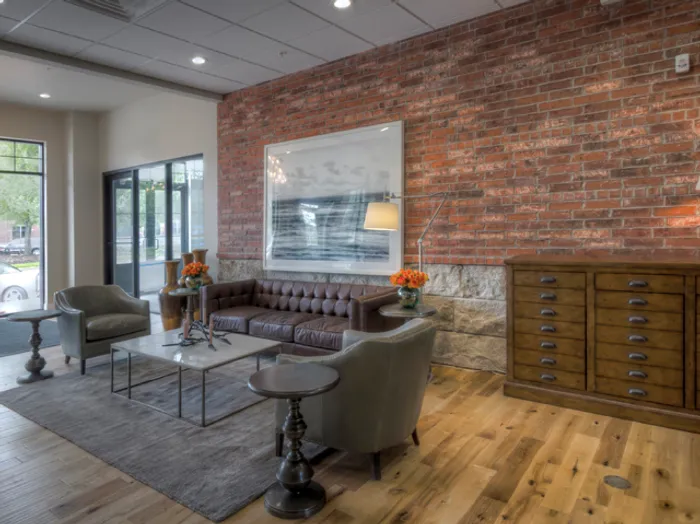 Stylish lobby at Chesterfield Lofts in Springfield, MO, featuring an exposed red brick and stone accent wall. The modern apartment lounge includes a tufted brown leather sofa, two gray armchairs, and a sleek glass-top coffee table on a soft gray area rug. A large framed black-and-white ocean photograph hangs as a focal point, while decorative elements such as orange floral arrangements, elegant table lamps, and a wooden sideboard enhance the inviting ambiance. Natural light streams in through large windows, highlighting the hardwood flooring and sophisticated design of this upscale Springfield, Missouri apartment community.