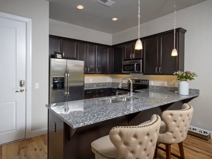 Modern kitchen at Chesterfield Lofts in Springfield, MO, featuring a large granite island with bar seating, stainless steel appliances, and dark wood cabinetry. Pendant lighting and open shelving add a sleek, contemporary touch to this high-end apartment kitchen.