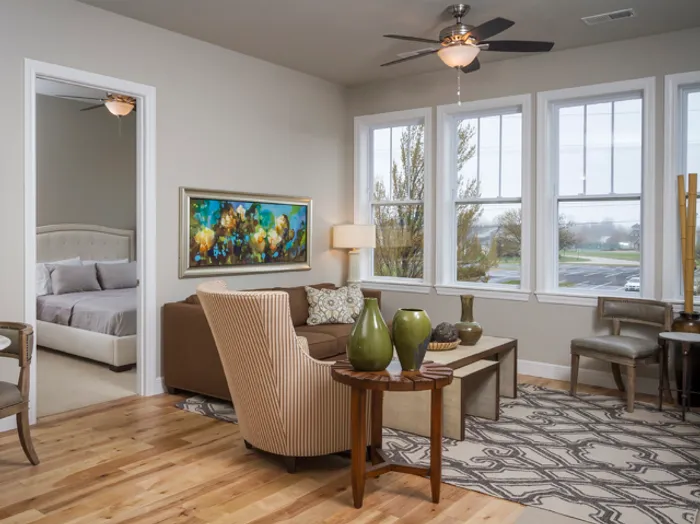 Bright and airy living room at Chesterfield Lofts in Springfield, MO, featuring large windows, a ceiling fan, and elegant furniture including a brown sofa, accent chairs, and stylish decor. A doorway leads to a modern bedroom with a tufted headboard and neutral-toned bedding, enhancing the luxury apartment&rsquo;s open design.