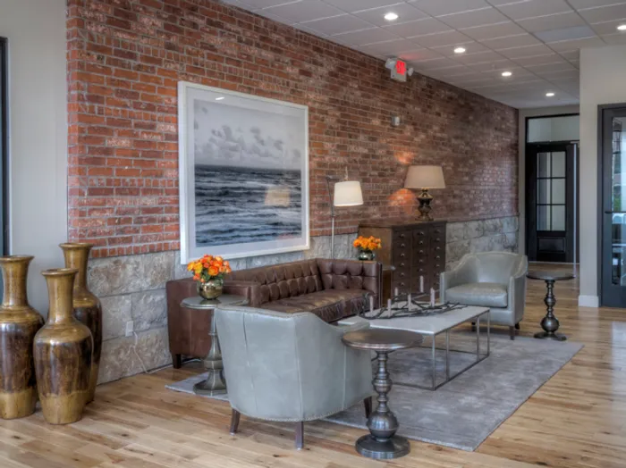 modern lounge area at Chesterfield Lofts in Springfield, MO, featuring an exposed red brick and stone accent wall. The upscale apartment lobby includes a tufted brown leather sofa, two sleek gray armchairs, and a glass-top coffee table set on a neutral-toned area rug. A large black-and-white ocean photograph adds a contemporary touch, while stylish d&eacute;cor elements such as table lamps, orange floral arrangements, and bronze vases enhance the inviting atmosphere. Hardwood flooring and recessed ceiling lighting contribute to the luxurious, industrial-chic aesthetic of this Springfield, Missouri apartment community.