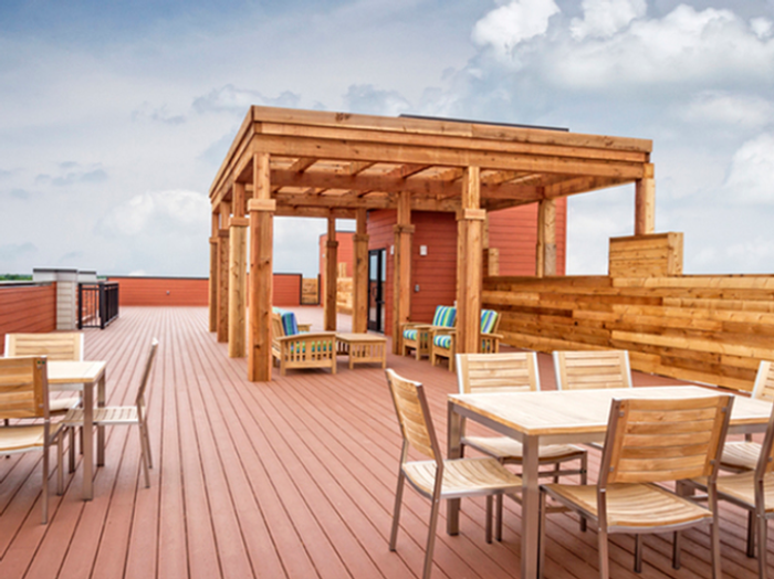 Expansive rooftop patio at Chesterfield Lofts in Springfield, MO, featuring a modern wooden pergola with comfortable lounge seating. The spacious outdoor deck includes multiple wooden dining tables with chairs, ideal for social gatherings or relaxing under the open sky. A warm wood-paneled privacy wall and sleek metal railing enclose the area, offering a stylish and inviting atmosphere. The scenic rooftop provides residents with a premium outdoor living space, perfect for enjoying fresh air and city views in this upscale Springfield, Missouri apartment community.