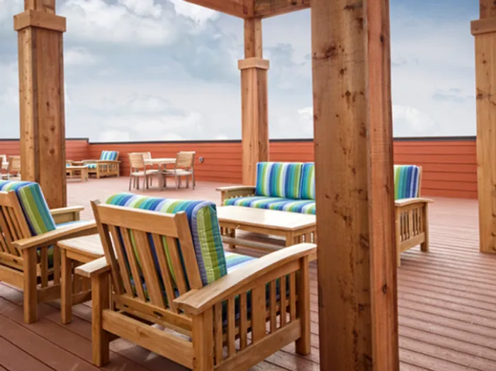 Close-up view of the rooftop patio at Chesterfield Lofts in Springfield, MO, showcasing a stylish wooden pergola with comfortable outdoor seating. The seating area features wooden chairs and sofas with vibrant blue and green striped cushions, creating a relaxing and inviting atmosphere. In the background, additional wooden dining tables and chairs are arranged on the spacious deck, surrounded by a modern red-paneled privacy wall. This upscale outdoor amenity provides residents with a scenic and comfortable space to unwind, socialize, and enjoy the open-air ambiance of this premium Springfield, Missouri apartment community.