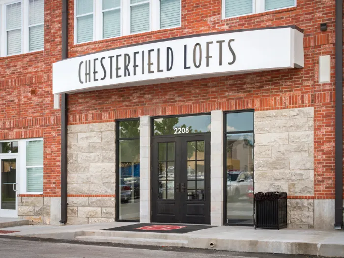 Front entrance of Chesterfield Lofts in Springfield, MO, featuring a black double door with glass panels, large street-facing windows, and a prominent white sign with bold black lettering above the entrance. The building exterior includes red brick and light stonework.