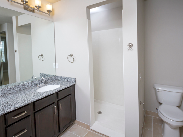 Modern bathroom at Chesterfield Lofts in Springfield, MO, featuring a sleek granite vanity with dark wood cabinetry, a large mirror, and elegant light fixtures. The space includes a walk-in shower with a white surround, a built-in towel hook, and a standard toilet. Neutral-toned tile flooring and soft gray walls create a clean and contemporary design in this upscale apartment home.