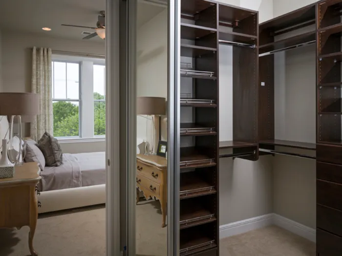 Spacious walk-in closet at Chesterfield Lofts in Springfield, MO, featuring custom dark wood shelving and mirrored sliding doors. The closet connects to a modern bedroom with a white tufted bed and large windows, offering ample storage space in this upscale Springfield, Missouri apartment.