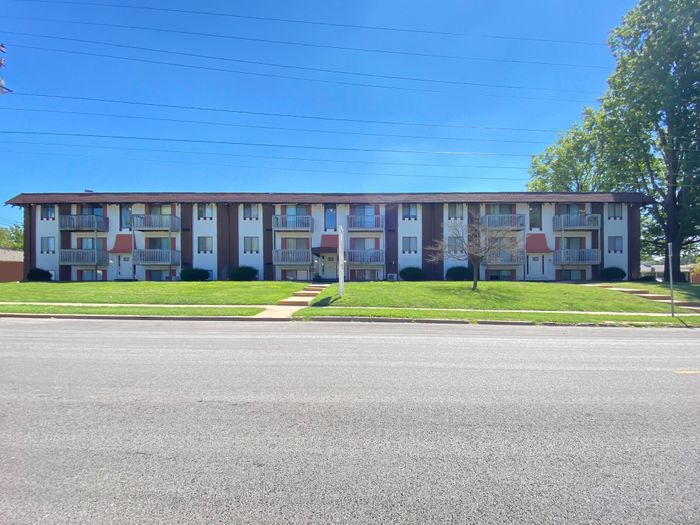 Front exterior of El Chaparral Apartments &ndash; Wide-angle street view of a three-story red brick apartment complex with white balconies, red awnings, and a large green lawn under a bright blue sky.