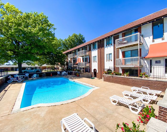 Swimming pool area &ndash; Outdoor pool surrounded by lounge chairs and shaded picnic tables, with a three-story apartment building and mature trees providing shade on a sunny day.