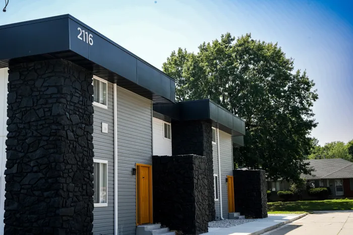 Modern apartment building with black stone columns, gray siding, and bright yellow entry doors. Address number 2116 is visible on the overhang.