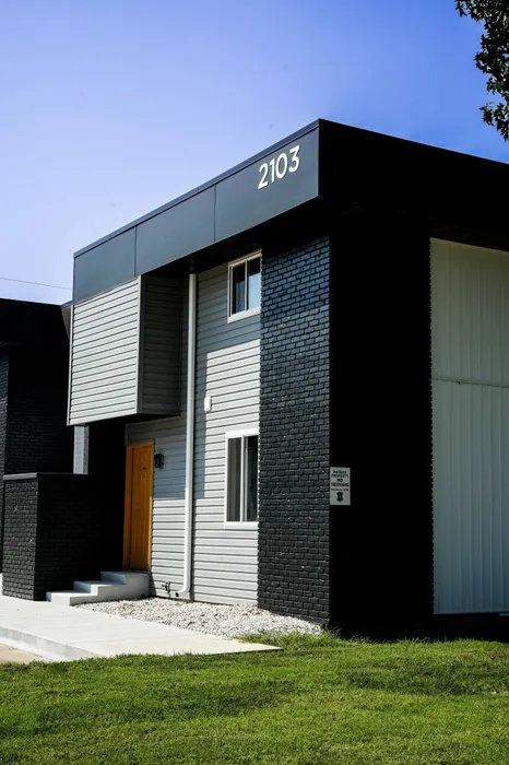 Corner unit of apartment building numbered 2103. Features gray siding, black brickwork, and a yellow door. 'Private property, no trespassing' sign visible.