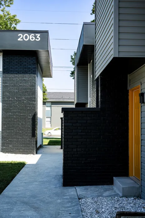 View between two apartment buildings with gray siding and yellow doors. Unit number 2063 visible. A concrete path runs between the buildings.